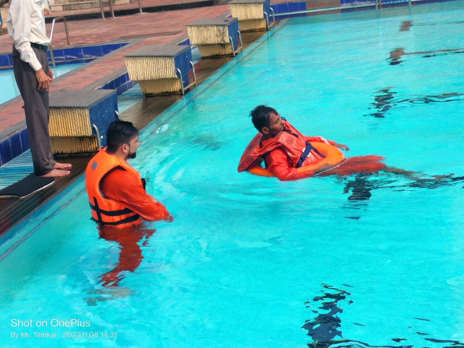 Two trainees in orange life jackets practising floating in a swimming pool while an instructor watches from the side