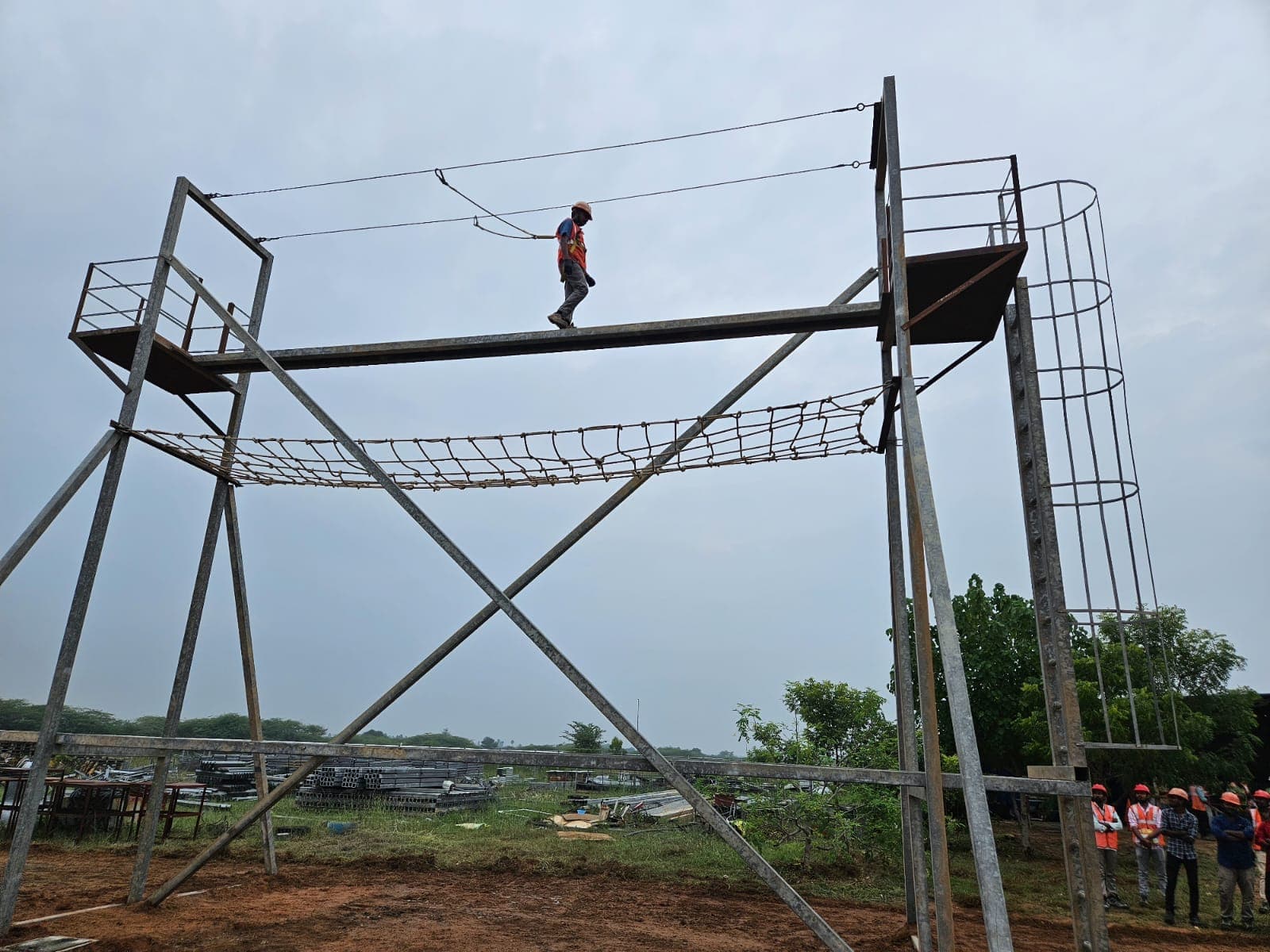 Trainee walking the top beam of a work-at-height training gantry, clipped to the overhead horizontal lifeline, with a group watching below