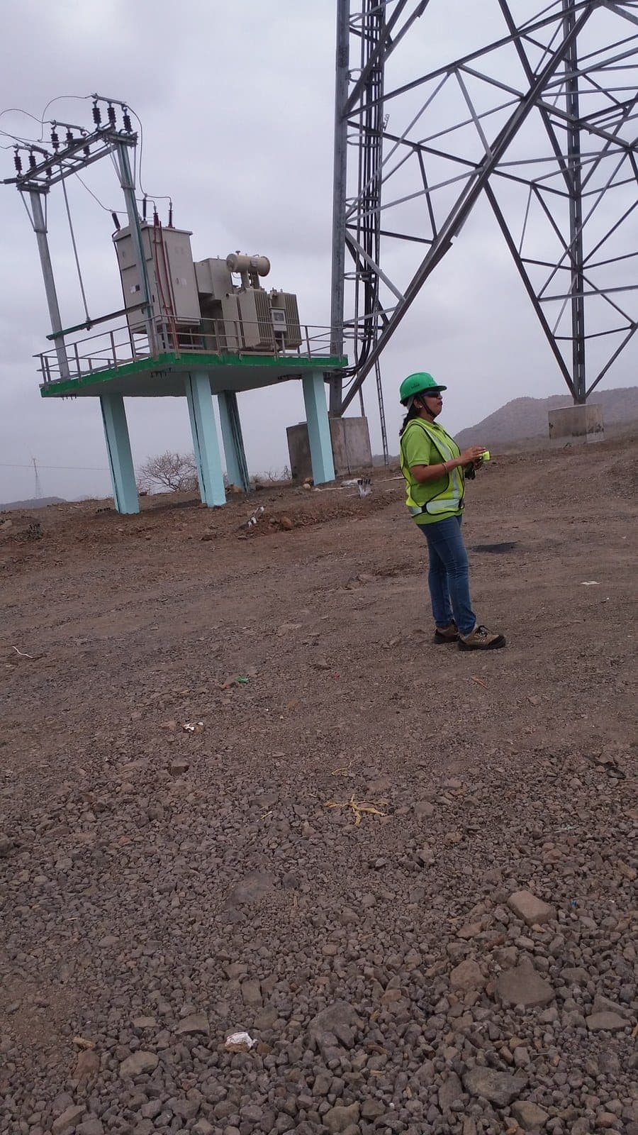 Person in a green helmet and high-visibility vest standing near a transformer and a transmission tower