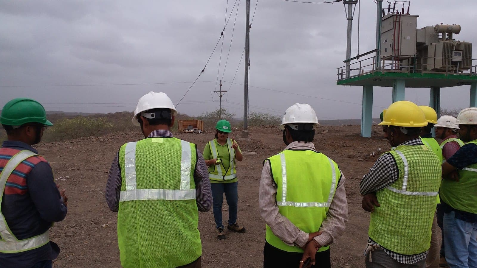 Workers in helmets and high-visibility vests listening to a site briefing beside an outdoor transformer