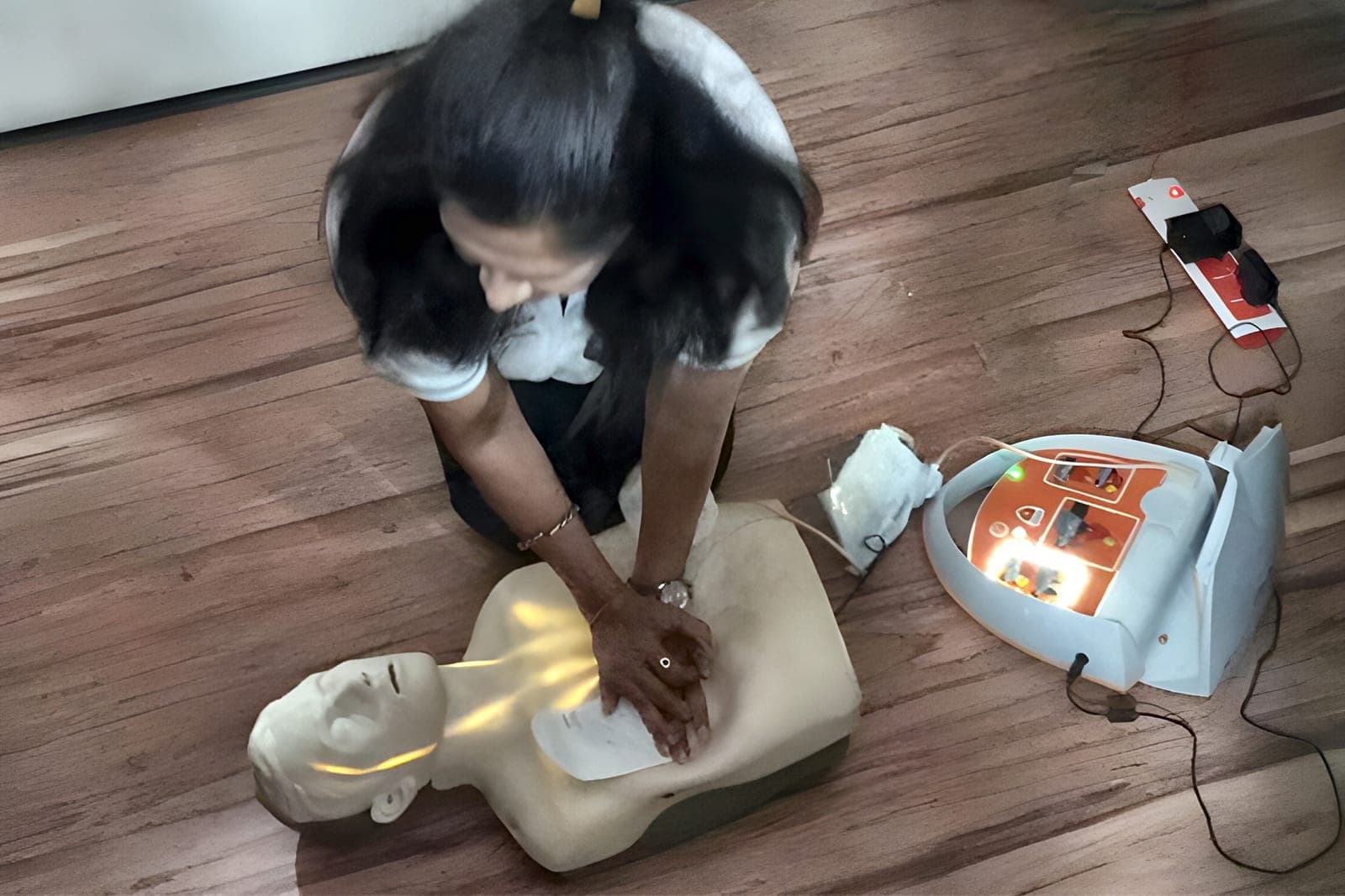 Trainee practising CPR chest compressions on a manikin, with an AED training unit beside her