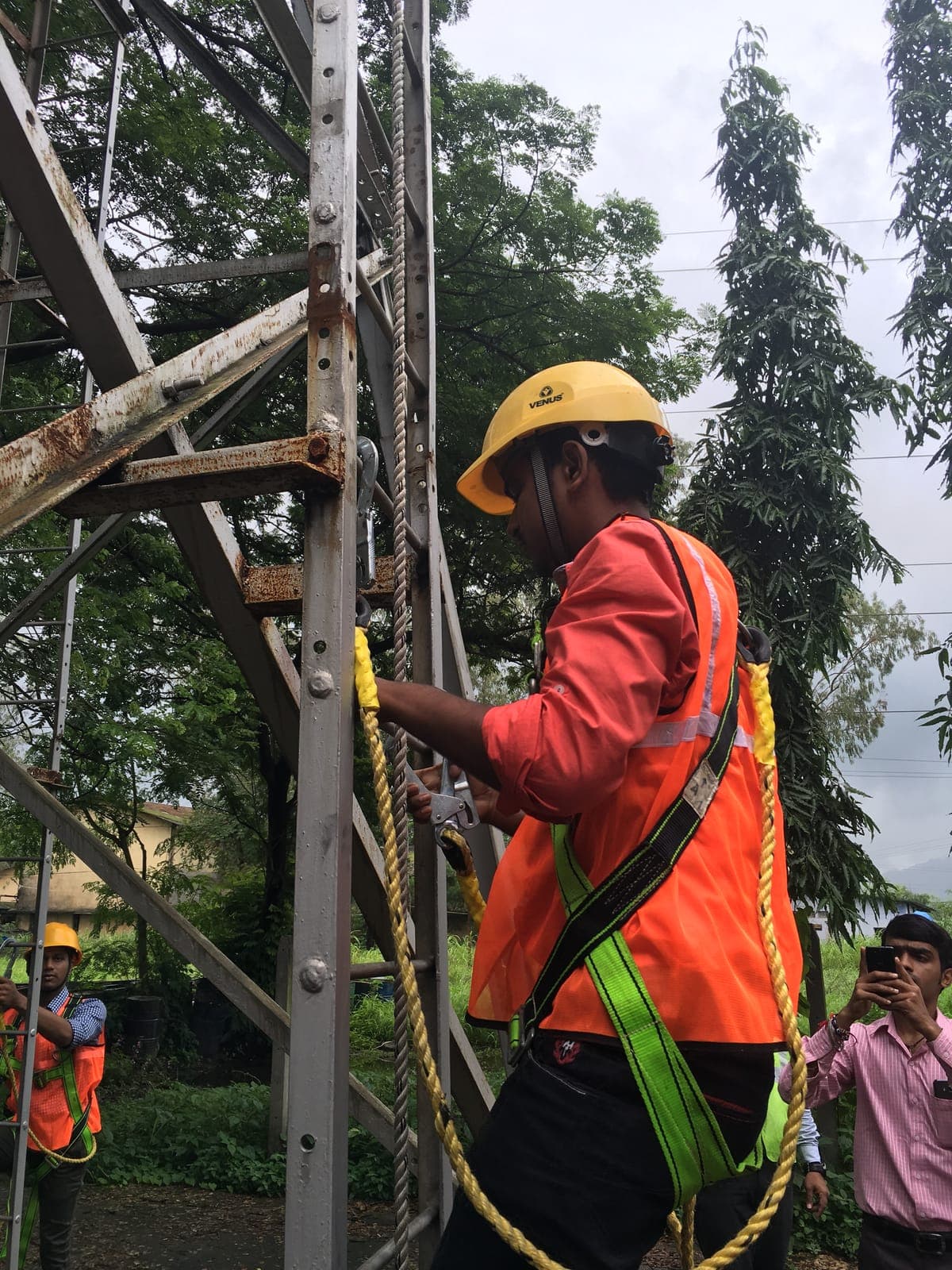 Trainee climbing a steel training tower in a hard hat, high-visibility vest and full-body harness, keeping one lanyard hook clipped on above while he moves the other